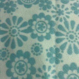 May include: Close-up of a white fabric with a repeating pattern of light blue flowers. The flowers have a retro, daisy-like design, with detailed petal shapes and central circular patterns.
