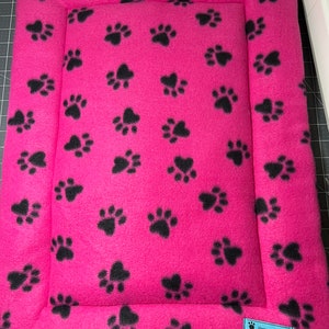 May include: A pink pet bed with a black paw print pattern. The bed has a raised edge and a label that says "Crafted To Comfort".