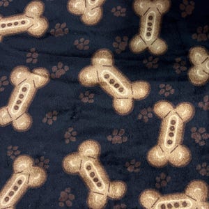 May include: Dark blue fabric with a repeating pattern of tan dog bones and paw prints. The dog bones have a detailed design with four small circles in the center. The paw prints are a solid tan color.