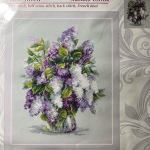 May include: A Riolis 1447 cross-stitch kit titled Gentle Lilac, featuring a bouquet of purple and white lilac flowers in a glass vase. The package includes stranded cotton and instructions for cross-stitch, half cross-stitch, back stitch, and French knot techniques, measuring 9 1/2 inches by 11 3/4 inches.
