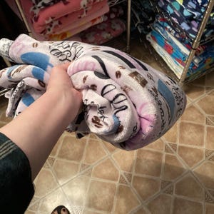 May include: A rolled-up plush blanket featuring a coffee-themed pattern in shades of pink, blue, and brown. The blanket is held in a person's hand, with other blankets visible on shelves in the background.