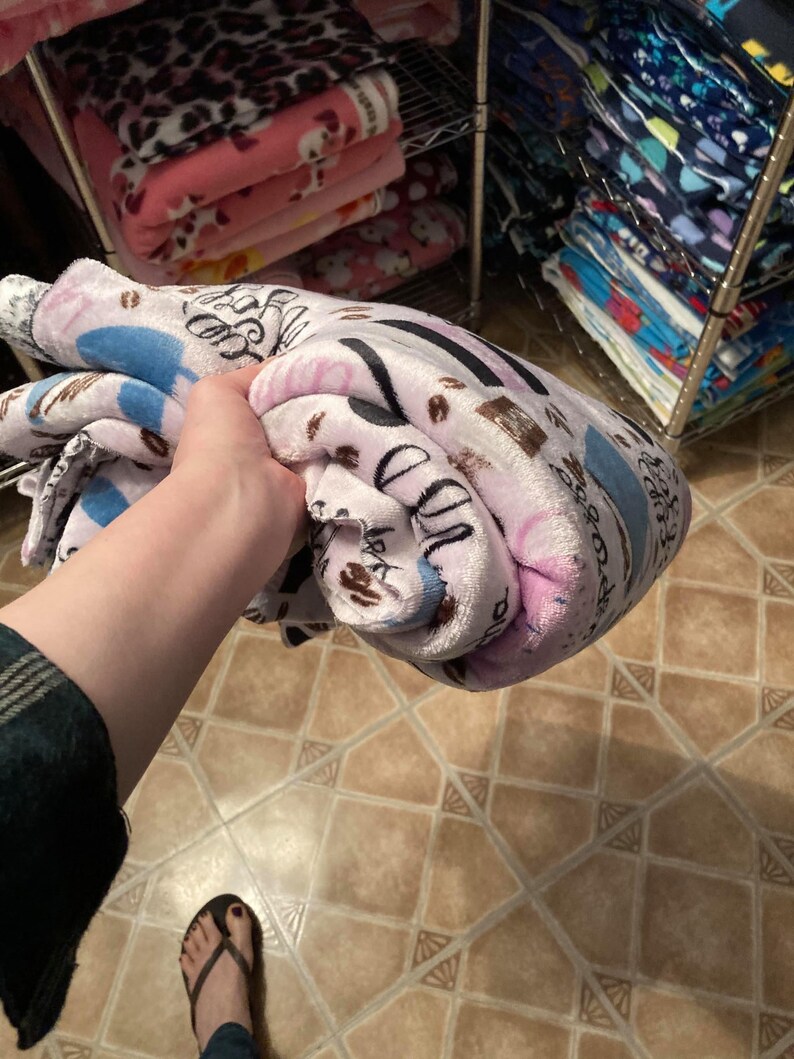May include: A rolled-up plush blanket featuring a coffee-themed pattern in shades of pink, blue, and brown. The blanket is held in a person's hand, with other blankets visible on shelves in the background.