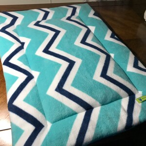 May include: A turquoise and white chevron patterned pet bed with navy blue trim. The bed has a sewn-in border and a green label with the text "Handmade by Gigi".