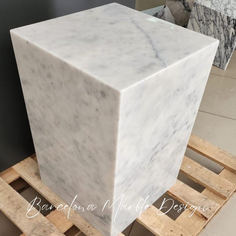 May include: A white marble cube stool with a smooth, polished surface. The stool is sitting on a wooden pallet.