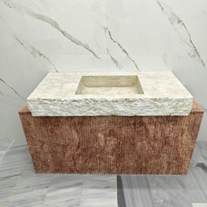 May include: A rectangular stone sink featuring a beige, textured, rough-hewn top surface with a central basin. The base of the sink is constructed from a solid, reddish-brown stone block with a vertical, linear-textured finish on the front face.