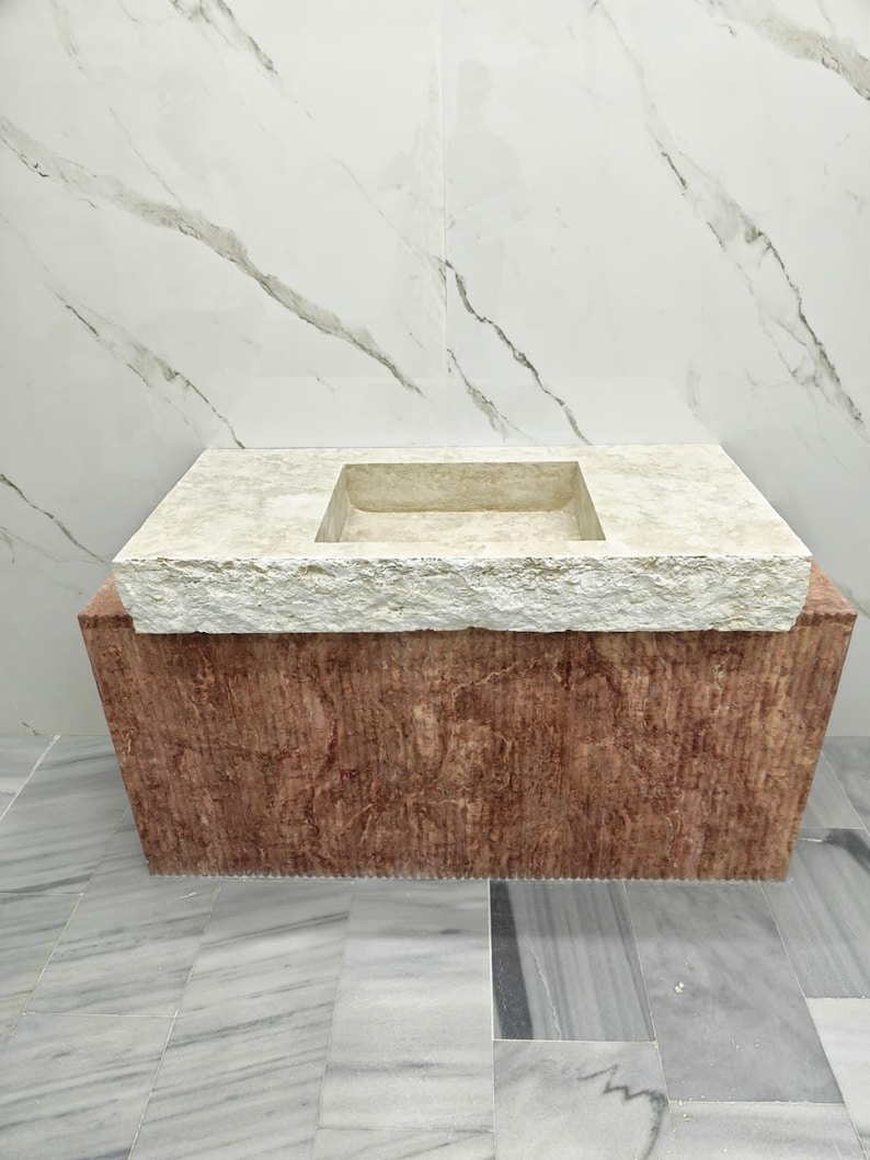 May include: A rectangular stone sink featuring a beige, textured, rough-hewn top surface with a central basin. The base of the sink is constructed from a solid, reddish-brown stone block with a vertical, linear-textured finish on the front face.
