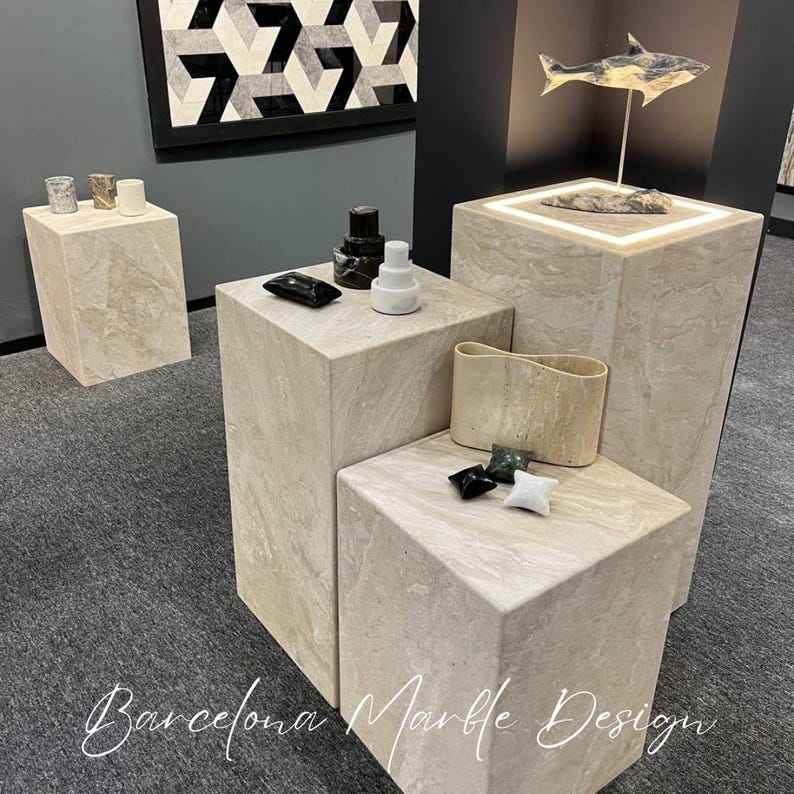 Puede incluir: Cuatro expositores de m&aacute;rmol beige de diferentes tama&ntilde;os, con un dise&ntilde;o de azulejos geom&eacute;tricos en blanco y negro en la pared detr&aacute;s de ellos. Los expositores est&aacute;n decorados con peque&ntilde;os objetos decorativos, incluyendo una escultura de un pez en blanco y negro.
