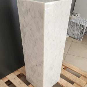 May include: A white marble pedestal with a smooth, polished surface. The pedestal is rectangular in shape and has a square base. It is sitting on a wooden pallet.