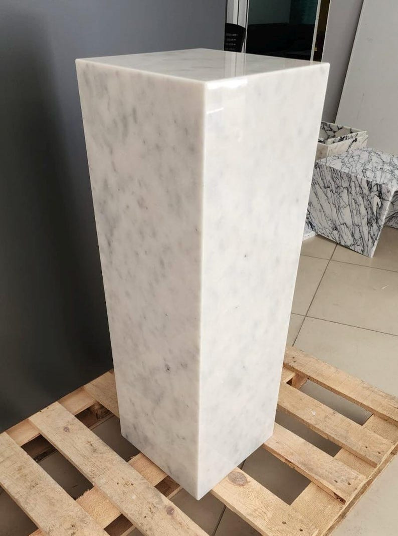 May include: A white marble pedestal with a smooth, polished surface. The pedestal is rectangular in shape and has a square base. It is sitting on a wooden pallet.