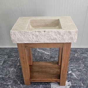 May include: A rectangular beige stone sink with a rough, textured front edge, mounted on a wooden vanity base. The vanity features four vertical legs and a lower shelf, constructed from light brown wood with a visible grain pattern throughout.