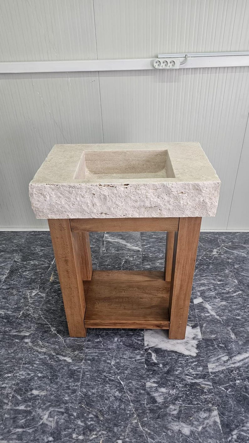 May include: A rectangular beige stone sink with a rough, textured front edge, mounted on a wooden vanity base. The vanity features four vertical legs and a lower shelf, constructed from light brown wood with a visible grain pattern throughout.