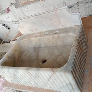 May include: A white marble sink with a decorative fluted base and a backsplash. The sink has three holes for faucets and a drain.