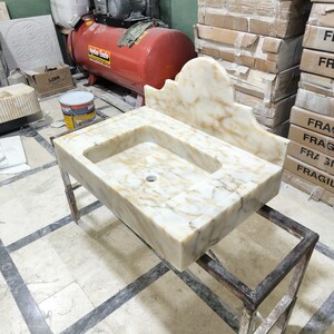 Puede incluir: Un lavabo rectangular de m&aacute;rmol con un protector contra salpicaduras incorporado, con un patr&oacute;n veteado beige claro y marr&oacute;n. El lavabo est&aacute; montado sobre un soporte met&aacute;lico. El fondo incluye cajas y equipos industriales.