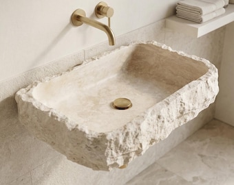 May include: A rectangular wall-mounted bathroom sink carved from beige natural stone with a rough, chiseled exterior texture and a smooth interior basin. It features a brushed gold faucet and a matching circular drain plug.