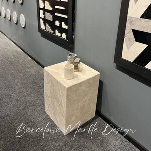 Puede incluir: Un pedestal cuadrado de m&aacute;rmol beige claro con una superficie texturizada. Dos peque&ntilde;as tazas de cer&aacute;mica blanca y un objeto dorado est&aacute;n encima del pedestal.