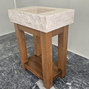 May include: A rectangular beige travertine stone sink basin with a rough, natural edge texture, mounted on a sturdy wooden vanity stand. The vanity features four thick vertical legs and a lower shelf, constructed from medium-toned brown wood.