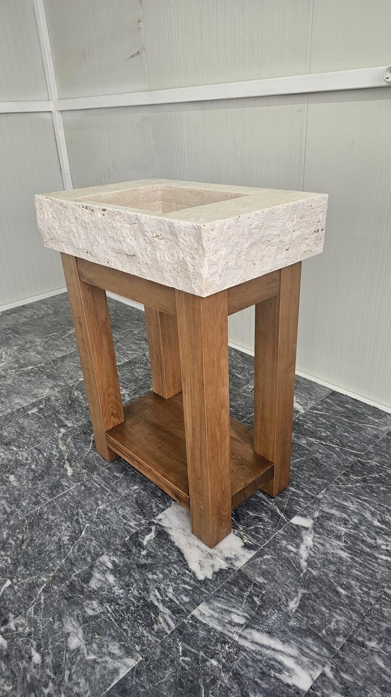 May include: A rectangular beige travertine stone sink basin with a rough, natural edge texture, mounted on a sturdy wooden vanity stand. The vanity features four thick vertical legs and a lower shelf, constructed from medium-toned brown wood.