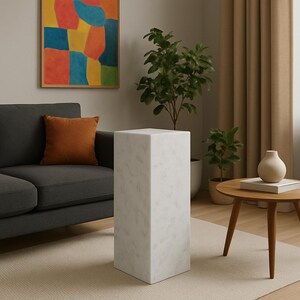 May include: A white marble pedestal with a square top and a smooth, polished finish. The pedestal is standing on a light beige rug in a living room setting.