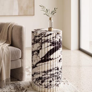 May include: A cylindrical side table with a marble design. The table features a white base with black veining and a fluted texture. A small glass vase with a gold base and a sprig of greenery sits on top. The table is approximately 20 inches tall.