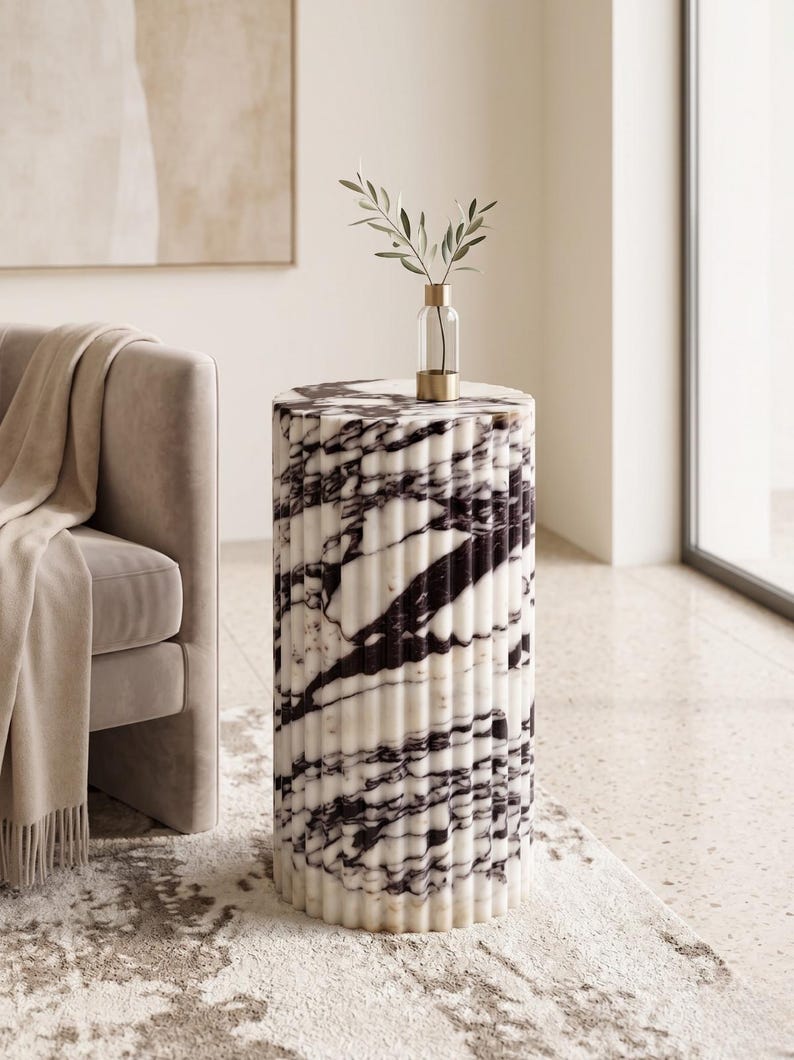May include: A cylindrical side table with a marble design. The table features a white base with black veining and a fluted texture. A small glass vase with a gold base and a sprig of greenery sits on top. The table is approximately 20 inches tall.