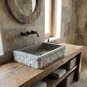 Puede incluir: Un tocador de baño rústico con un lavabo rectangular de piedra. El tocador está hecho de madera reciclada con dos cestas de mimbre y estantes. Un espejo redondo de madera y un grifo de bronce completan el diseño.