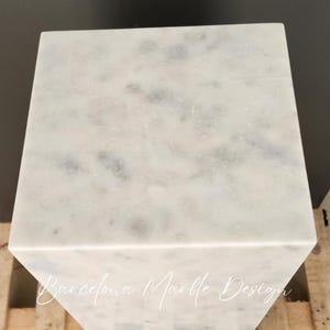 May include: White marble pedestal with a gray veining pattern. The pedestal is square and has a smooth, polished surface. The text "Barcelona Marble Design" is visible on the side of the pedestal.