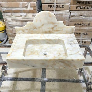Puede incluir: Un lavabo rectangular de m&aacute;rmol con un respaldo curvo. El lavabo es de color blanquecino con vetas marrones y un orificio de desag&uuml;e central. Se exhibe sobre un marco de metal, con cajas etiquetadas como "FR&Aacute;GIL" en el fondo.