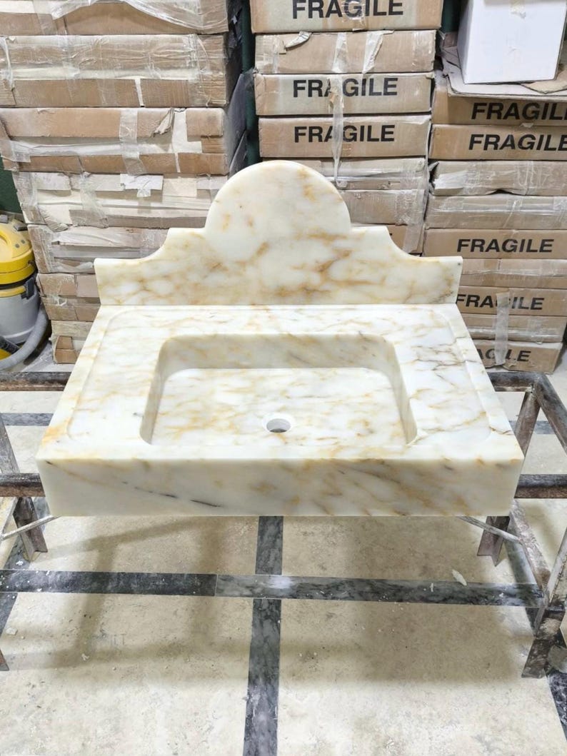 Puede incluir: Un lavabo rectangular de m&aacute;rmol con un respaldo curvo. El lavabo es de color blanquecino con vetas marrones y un orificio de desag&uuml;e central. Se exhibe sobre un marco de metal, con cajas etiquetadas como "FR&Aacute;GIL" en el fondo.