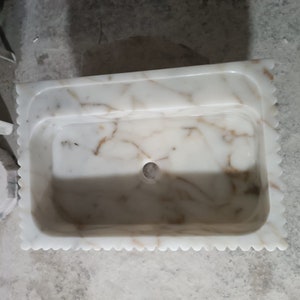 May include: White marble sink with a scalloped edge and a drain hole in the center. The sink has a light brown veining pattern.