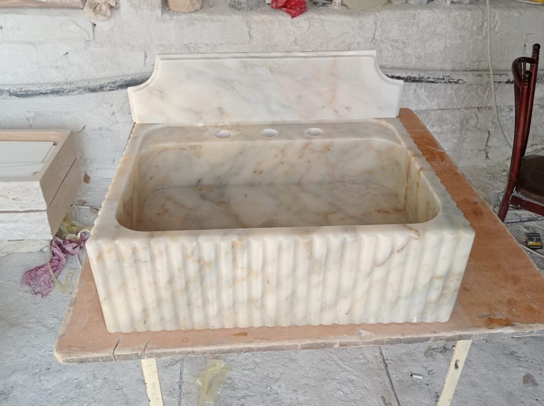 May include: A white marble sink with a decorative backsplash and a fluted base. The sink has three holes for faucets.