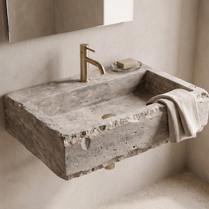 May include: A rectangular wall-mounted bathroom sink carved from natural grey stone with a rough, textured exterior. It features a brushed gold tap, a matching gold drain, and a folded beige cloth draped over the front edge of the basin.