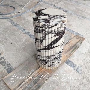 May include: A cylindrical marble pedestal with a fluted design. The pedestal is white with black veining and sits on a wooden plank. The text "Barcelona Marble Design" is at the bottom.