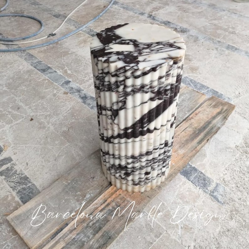 May include: A cylindrical marble pedestal with a fluted design. The pedestal is white with black veining and sits on a wooden plank. The text "Barcelona Marble Design" is at the bottom.