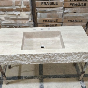 May include: A rectangular beige travertine stone bathroom vanity top featuring a central integrated rectangular basin, three faucet holes, and a rough, chiseled natural stone front edge. The vanity is supported by a simple metal frame base.