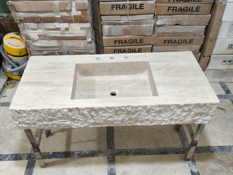 May include: A rectangular beige travertine stone bathroom vanity top featuring a central integrated rectangular basin, three faucet holes, and a rough, chiseled natural stone front edge. The vanity is supported by a simple metal frame base.
