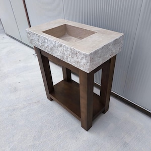 May include: A rectangular stone sink with a rough, textured exterior and a smooth basin, mounted on a dark brown wooden stand featuring four straight legs and a lower shelf. The sink has a light beige, natural stone finish with visible grain.