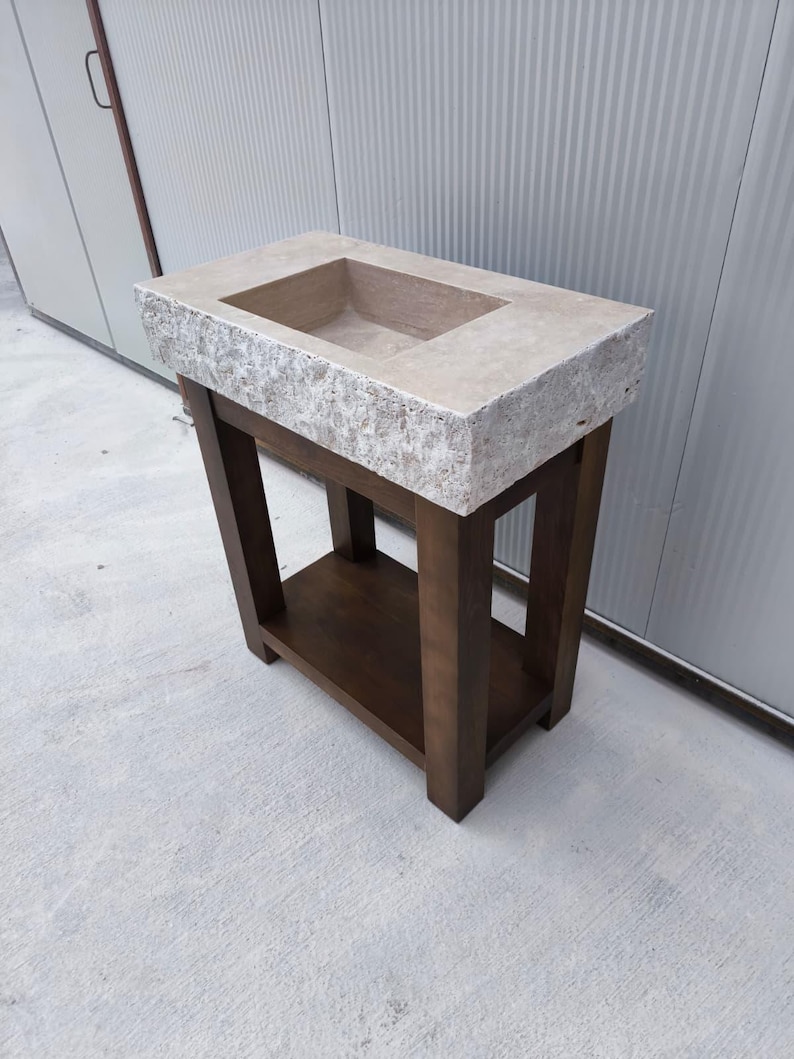 May include: A rectangular stone sink with a rough, textured exterior and a smooth basin, mounted on a dark brown wooden stand featuring four straight legs and a lower shelf. The sink has a light beige, natural stone finish with visible grain.
