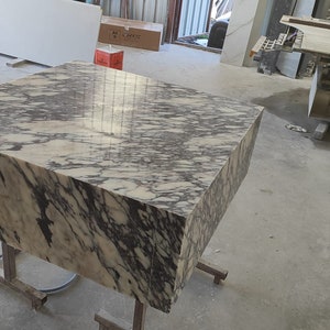 K&ouml;nnte beinhalten: Ein gro&szlig;er, quadratischer, wei&szlig;er Marmortisch mit schwarzen Adern. Der Tisch steht auf einem Metallgestell mit R&auml;dern. Der Tisch befindet sich in einer Werkstatt.