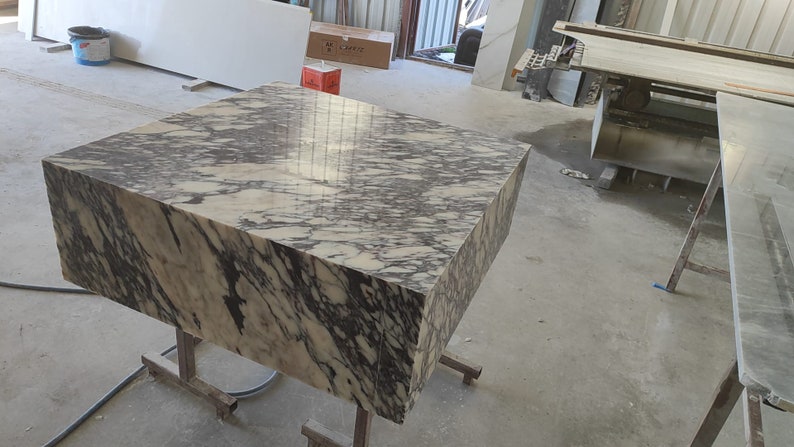 K&ouml;nnte beinhalten: Ein gro&szlig;er, quadratischer, wei&szlig;er Marmortisch mit schwarzen Adern. Der Tisch steht auf einem Metallgestell mit R&auml;dern. Der Tisch befindet sich in einer Werkstatt.