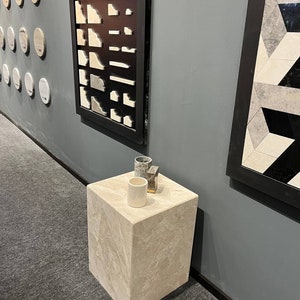 Puede incluir: Un pedestal cuadrado beige claro con tres peque&ntilde;os objetos decorativos en la parte superior. Los objetos son una taza de cer&aacute;mica gris, una taza de cer&aacute;mica blanca y un peque&ntilde;o objeto de metal gris y dorado. El pedestal est&aacute; sobre una alfombra gris.