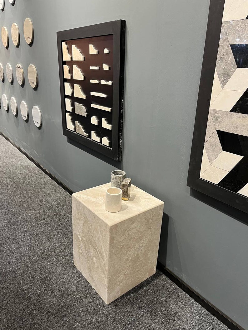 Puede incluir: Un pedestal cuadrado beige claro con tres peque&ntilde;os objetos decorativos en la parte superior. Los objetos son una taza de cer&aacute;mica gris, una taza de cer&aacute;mica blanca y un peque&ntilde;o objeto de metal gris y dorado. El pedestal est&aacute; sobre una alfombra gris.