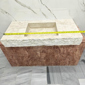 May include: A rectangular stone sink with a beige, textured top surface and a reddish-brown base. A yellow measuring tape is stretched across the top, indicating a length of approximately 47 inches from the left edge to the right edge.