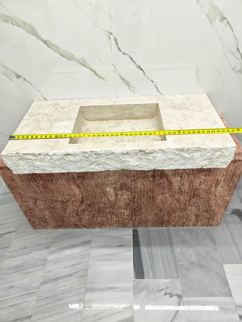 May include: A rectangular stone sink with a beige, textured top surface and a reddish-brown base. A yellow measuring tape is stretched across the top, indicating a length of approximately 47 inches from the left edge to the right edge.