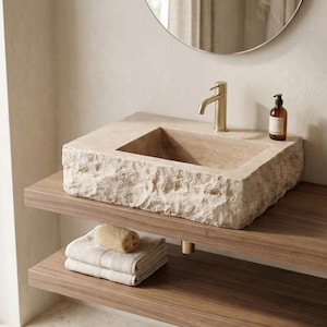 Puede incluir: Un lavabo rectangular con aspecto de piedra con un grifo de latón. El lavabo está sobre una estantería de madera oscura, con toallas dobladas y una esponja debajo. Un espejo redondo cuelga sobre el lavabo y una botella de jabón está en la encimera.