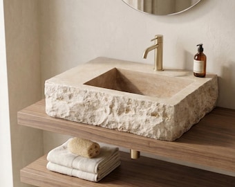 May include: A stone-like rectangular bathroom sink with a brass faucet. The sink sits on a dark wood shelf, with folded towels and a sponge below. A round mirror hangs above the sink, and a bottle of soap sits on the counter.