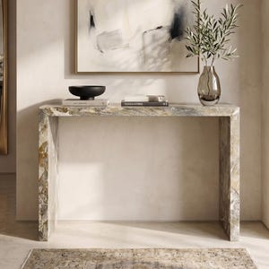 May include: A marble console table with a black bowl, books, and a glass vase with olive branches. The table is rectangular with a natural stone pattern. An abstract painting hangs above the table, and a rug is on the floor.