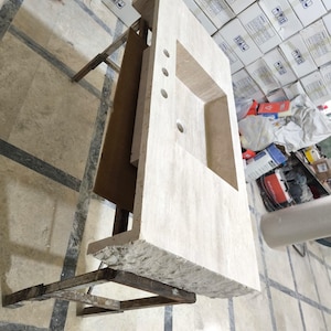 May include: A rectangular beige travertine vanity countertop featuring a single integrated rectangular sink basin, three pre-drilled faucet holes, and a rough, natural chiseled edge along the front side, supported by a metal frame structure.