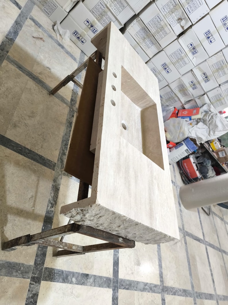 May include: A rectangular beige travertine vanity countertop featuring a single integrated rectangular sink basin, three pre-drilled faucet holes, and a rough, natural chiseled edge along the front side, supported by a metal frame structure.