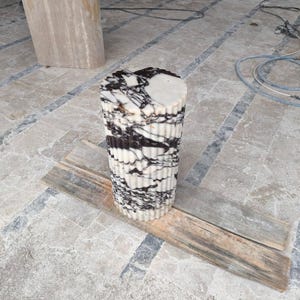 May include: A cylindrical marble accent table with a fluted design. The table features a white base with black veining and a flat top. The table is approximately 24 inches tall and is sitting on a wooden plank.