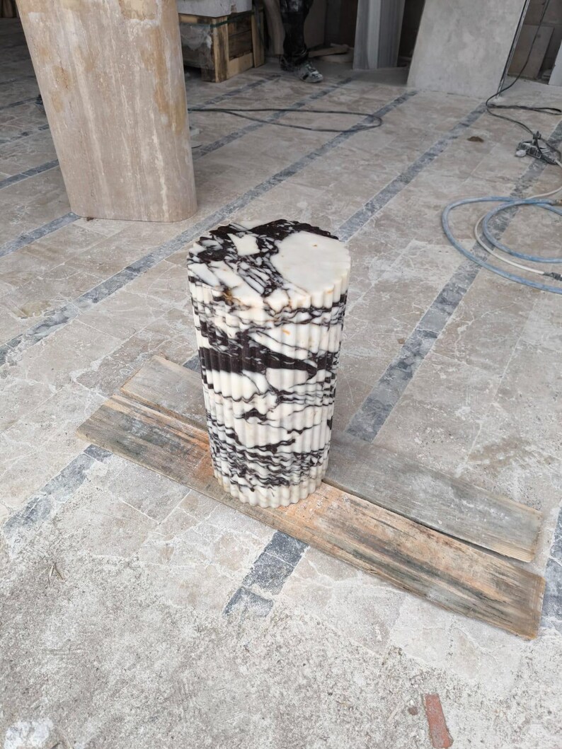 May include: A cylindrical marble accent table with a fluted design. The table features a white base with black veining and a flat top. The table is approximately 24 inches tall and is sitting on a wooden plank.